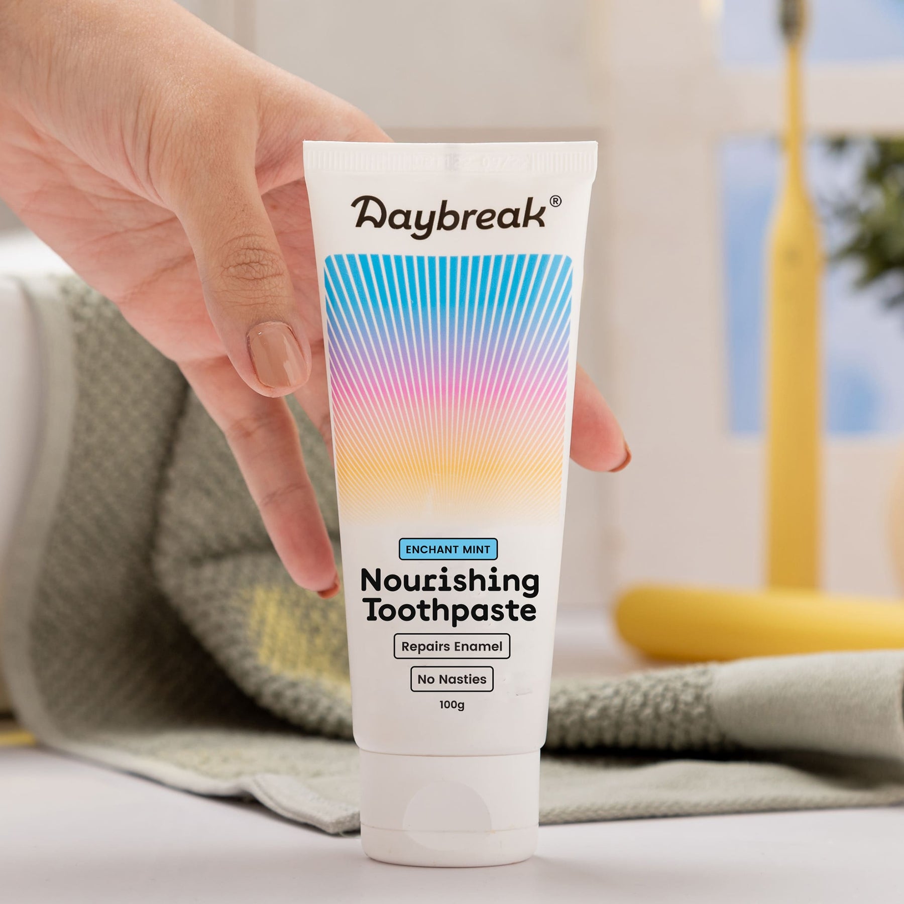 A hand reaching for the Daybreak Brighten Toothpaste sitting on a bathroom shelf